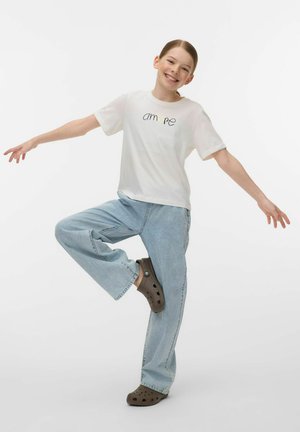 Young person smiling with arms outstretched, balancing on one leg, wearing white t-shirt, light blue jeans, and brown slip-on shoes.