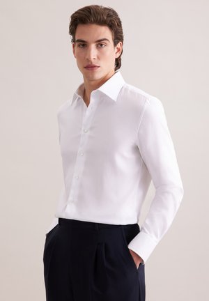 Młody mężczyzna w białej koszuli na guziki wpuszczonej w czarne plisowane spodnie, stojący z rękami w kieszeniach na jednolitym tle.