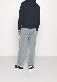 Sudadera azul marino con un bolsillo canguro, combinada con jeans holgados de color gris claro. Los zapatos son negros con detalles en blanco. La textura parece suave y casual.