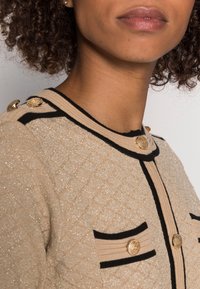 Beige strukturerad kavaj med guldfärgade knappar, svart kant och fickor. Tyget har en subtil lyster och ett quiltat mönster.