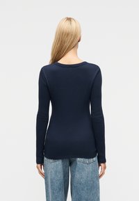 Γυναίκα με μακριά ξανθά μαλλιά, φαίνεται από πίσω, φοράει μπλε σκούρο ριπ μακρυμάνικο πουλόβερ και ανοιχτά μπλε τζιν ψηλόμεσα, πάνω σε απλό φόντο.