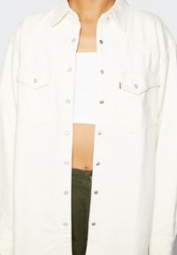 Persona que lleva una chaqueta de mezclilla blanca desabotonada sobre una camiseta corta blanca y pantalones verde oliva, mostrando el abdomen con el ombligo.