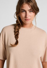 Jonge vrouw met een losse vlecht over haar linkerschouder, gekleed in een beige T-shirt met korte mouwen, die recht in de camera kijkt met een neutrale uitstraling.