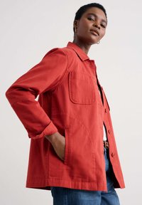 Veste en coton rouge avec un col, fermeture à boutons, poches à rabat et une coupe légèrement décontractée. Coutures visibles et tissu texturé.