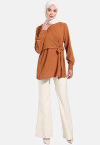Haut marron à manches longues et lien sur le côté, associé à un pantalon blanc évasé. Le tissu semble doux avec une texture lisse.