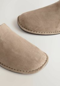 Gros plan de chaussures beiges en daim à enfiler avec des bouts carrés arrondis et des coutures visibles le long des bords sur une surface claire.