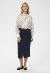 Blusa blanca con estampado floral y cuello con lazo, combinada con una falda midi de mezclilla oscura con una abertura frontal. Zapatos planos tipo ballet negros completan el conjunto.