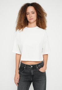 Fehér rövid fazonú póló, rövid, széles ujjakkal és kerek nyakkivágással, sötétszürke, magas derekú farmernadrággal párosítva, elülső zsebekkel.