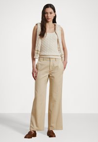 Pantaloni bej cu picioare largi, cu talie înaltă și textură fină, asortați cu un top fără mâneci din tricot cu model de cablu de culoare crem și un pulover aruncat peste umeri.