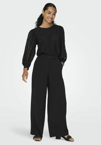 Zwarte kanten blouse met lange mouwen en pofmouwen, gecombineerd met hoge zwarte wijde broek en zwarte sandaaltjes met bandjes.
