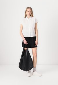 Camicetta bianca a maniche corte e colletto con ricami floreali, abbinata a pantaloni corti neri. Tiene una grande borsa a tracolla in pelle nera. Sneakers bianche.