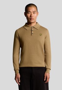 Brun långärmad polotröja med krage, tre knappar och en gul broderad logotyp på vänster bröst. Mjuk textur, avslappnad passform.