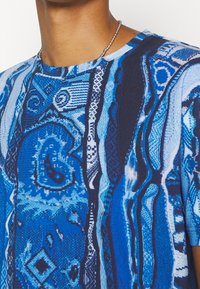 Nära bild av en person som bär en blå mönstrad skjorta med abstrakta och geometriska mönster, samt ett halsband med silverpärlor.