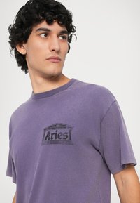 Tricou din bumbac violet cu mâneci scurte, având un design imprimat în negru pe piept. Textură moale cu o croială lejeră.