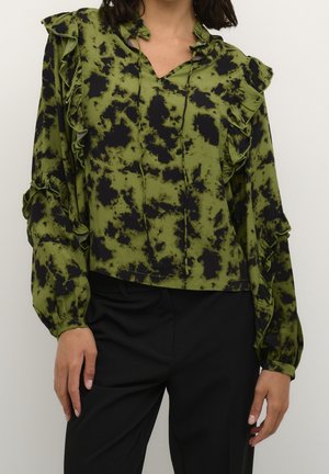 Kobieta ubrane w zieloną i czarną bluzkę w cętki z falbanami i długimi rękawami, w połączeniu z czarnymi spodniami, stojąca na jednolitym tle.