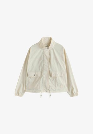 Lekka jasnobeżowa kurtka z wysokim kołnierzem, z przodu zapięcie na napy, dwie kieszenie z patkami, elastyczne mankiety i regulowane sznurki w dolnym obszyciu.