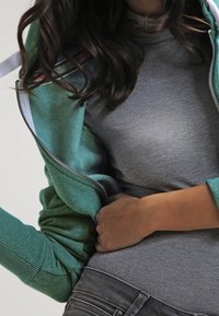 Sudadera verde con cremallera y mangas texturizadas, sobre una camiseta gris ajustada. Incluye un detalle de rayas sutil en la capucha y puños acanalados.