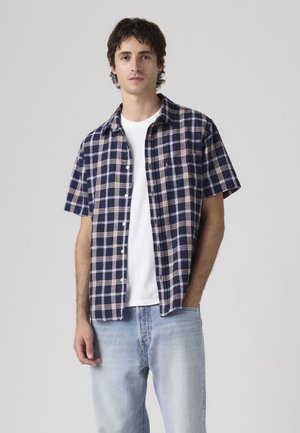 Bărbat tânăr purtând o cămașă cu mâneci scurte, în carouri bleumarin și bej, peste un tricou alb, și blugi deschiși la culoare, stând cu o mână în buzunar.