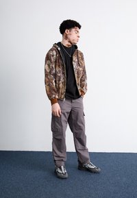 Giacca zip-up mimetica con tonalità di marrone e grigio, t-shirt nera, pantaloni cargo grigi e sneakers grigie con accenti neri.
