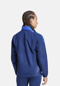 Veste coupe-vent marine avec col bleu et rayures, poignets élastiques et texture légère et douce. Vue arrière présentée.