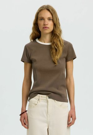 Jeune femme aux longs cheveux blonds ondulés, portant une chemise marron à manches courtes avec un col blanc et un pantalon couleur crème, debout devant un fond uni.