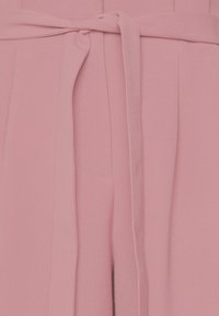 Rosa tygplagg med knutdetalj i midjan, slät yta och subtila vertikala veck; inkluderar sidofickor.