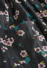 Tissu foncé avec imprimé floral blanc, violet et bleu, comprenant une poche cousue et un bouton en bois visible.