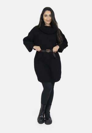 Frau trägt ein schwarzes gestricktes Pulloverkleid, schwarze Leggings und schwarze Schnürstiefel und justiert einen schwarzen Gürtel mit zwei goldenen Herzschnallen.