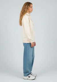 Felpa crema con tasca a marsupio, abbinata a jeans larghi di colore blu chiaro. Sneakers bianche con accenti neri. Sfondo neutro.