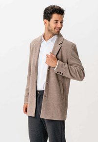 Mand justerer sin knapækket beige frakke over en hvid skjorte og mørkegrå bukser, mens han ser sidelæns og smiler mod en hvid baggrund.