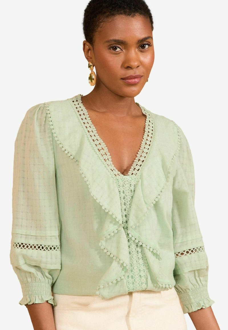 Svetlozelená blúzka s volánovým predným dielom, čipkovým lemovaním a vypchatými rukávmi. Obsahuje výstrih do V a jemné detaily pozdĺž rukávov.
