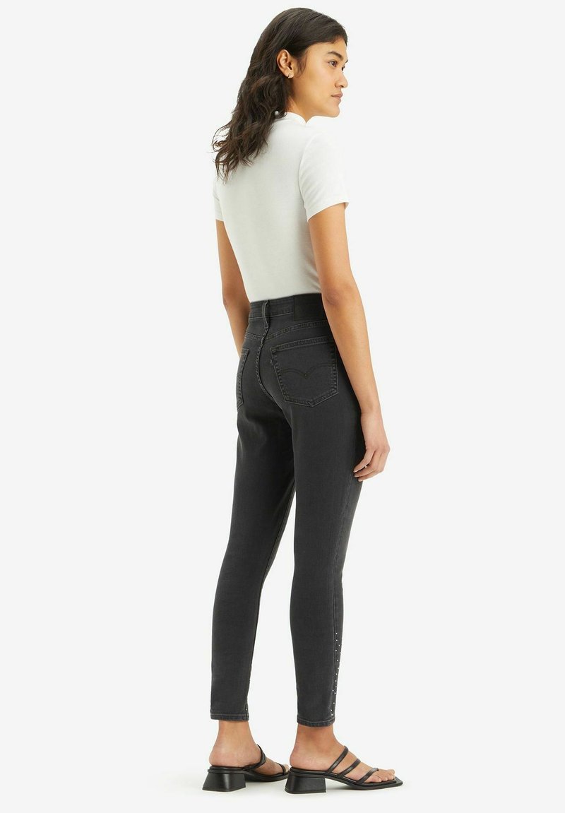 Hoch geschnittene schwarze Skinny-Jeans mit glatter Textur, ausgestattet mit Gesäßtaschen und seitlichen Verzierungen. Kombiniert mit einem weißen, figurbetonten Oberteil und schwarzen Sandalen.