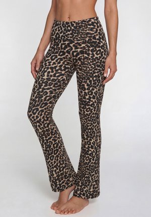 Frau trägt leopardengeprintete, hoch taillierte Leggings und steht barfuß vor einem schlichten, hellen Hintergrund.