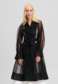 Sort mesh trenchcoat med dobbeltknaplukning, bindebælte i taljen og gennemsigtige lange ærmer. Har knapdetaljer og broderet logo.