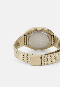 Montre en acier inoxydable doré avec un boîtier carré, un bracelet en maille et un dos argenté affichant le logo Adidas. Fermeture ajustable pour un bon ajustement.