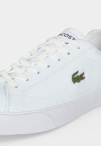 Λευκό αθλητικό παπούτσι Lacoste με λευκά κορδόνια, πράσινο λογότυπο κροκοδείλου στο πλάι και σήμανση Lacoste στη γλώσσα.
