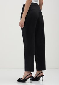 Femme debout portant un pantalon noir taille haute droit et des talons slingback noirs avec un détail en nœud, sur un fond blanc uni.