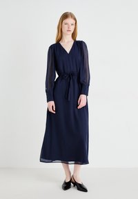 Navy blue maxi dress with long sheer sleeves, V-neckline, and a cinched waist. Features a ribbon tie and a flowing hemline.