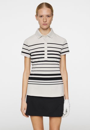 Polo shirt i lys beige med sorte vandrette striber, korte ærmer og en knapkrave. Styles med en sort nederdel og hvide handsker.