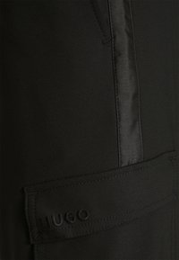 Pantalons noirs en tissu texturé, avec un panneau latéral en satin et une poche à rabat ornée du logo brodé "HUGO".