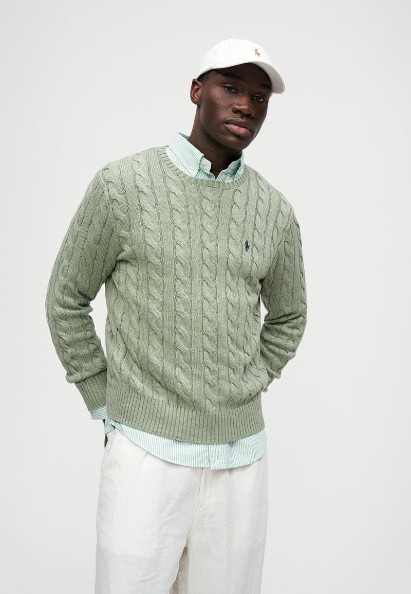 Suéter verde de punto trenzado con puños y dobladillo acanalados, superpuesto a una camisa de cuello a rayas azul claro y blanca, combinado con pantalones crema.