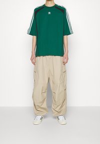 Person som bär en grön Adidas T-shirt med vita ränder, beige cargo-byxor och vita sneakers, stående mot en vit, enfärgad bakgrund.