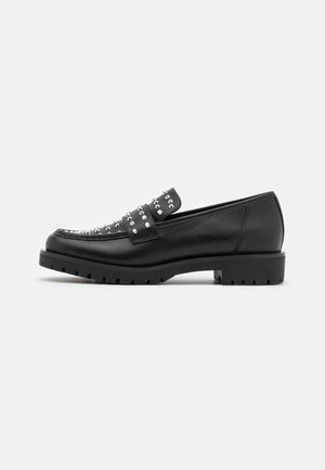 Mocassin en cuir noir avec semelle épaisse et clous argentés sur le dessus et la bride, vue de côté sur fond blanc.