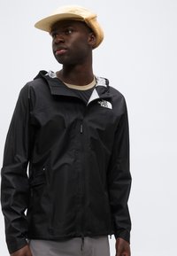 Άνδρας που φοράει μπεζ καπέλο τύπου bucket και μαύρο μπουφάν The North Face, κοιτάζοντας ελαφρώς προς το πλάι, με απλό φωτεινό φόντο.