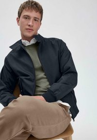 Zwart bomberjack voor mannen met een staande kraag, ritssluiting, ribgebreide manchetten, gedragen over een olijfgroen sweater en een wit shirt.