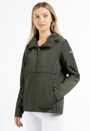 Femme portant une veste verte zippée avec capuche, debout avec une main dans la poche, regardant légèrement sur le côté sur fond blanc.