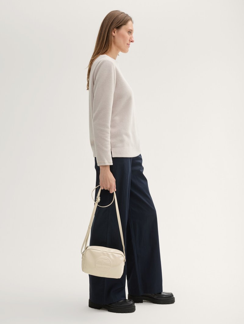 Femme en pull beige et pantalon large marine tenant un sac à bandoulière beige, debout de profil sur un fond uni.