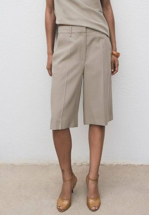 Pantalones cortos de corte recto en color beige con pliegues frontales, textura suave y presillas para cinturón, combinados con tacones abiertos marrones. Diseño simple y estructurado.