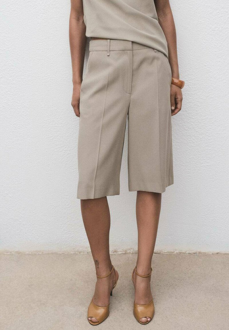 Beige skræddersyede shorts med frontfolder, glat tekstur og bæltestropper, parret med brune åbne hæle. Enkel, struktureret design.
