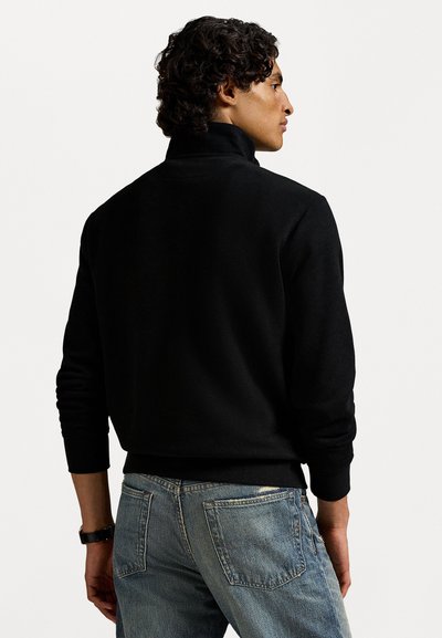 Mand med krøllet mørkt hår iført en sort langærmet trøje og slidte blå jeans, vendt let mod højre mod en ensfarvet baggrund.